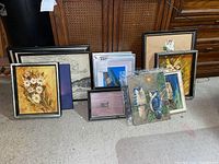 Overview photo showing a group of 14 diverse framed artworks including floral paintings, scenic sketches, and bird-themed pieces leaning against furniture on a carpet.