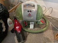Full view of the Bissell Littlegreen ProHeat carpet cleaner with green and white design alongside red and black Dirt Devil handheld vacuum on carpeted floor.