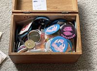 Open wooden box filled with assorted buttons, pins, and badges.