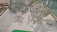Two clear crystal bear figurines and two clear crystal ashtrays shown on table, one bear larger with black eyes and nose, other smaller. Ashtrays are textured and faceted clear crystal.