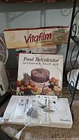 Box of Vitafilm food packaging film, Food Dehydrator accessory tray kit box, FoodSaver accessories packets and bags on carpet.
