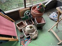 Overall assortment of garden items placed on green floor showing pots stacked, watering cans, decorative planter, and wind chimes.