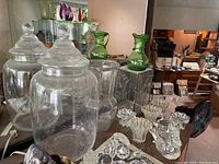 Photo showing multiple glass items on a table including two large glass apothecary jars with lids, green glass pitchers, several clear glass vases, and small ruffled edge candle holders.