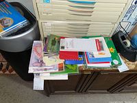 Stack of assorted notepads, greeting cards, and stationery on wooden cabinet next to paper storage trays and trash bin.