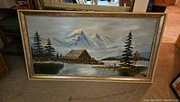 Full view of the framed landscape painting showing the mountain cabin, trees, and lake scene.