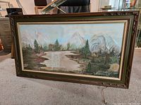Full view of original oil painting depicting mountain landscape with river, trees, greenery in wooden frame on carpeted floor.