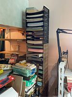 Tall stack of black plastic trays with papers and notebooks to the left of a wooden shelf with office supplies