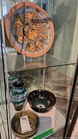 Glass display case showing brown and blue pottery plate, German-made vase with green and brown glaze, smaller dark glazed bowl, and another brown bowl.