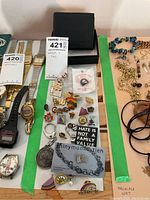 Overview of assorted pins laid out on a surface, with various designs including rainbow pride, political slogans, symbols, and metallic finishes. A black wallet box is also partially visible behind the pins.