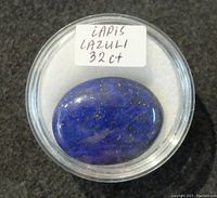 Top view of the oval lapis lazuli cabochon gemstone inside a clear plastic display container with a label showing 'LAPIS LAZULI 32ct'. The stone has a deep blue color with visible gold specks.