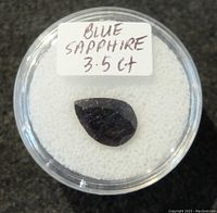 Clear round container holding a teardrop-shaped blue sapphire gemstone labeled 'BLUE SAPPHIRE 3.5 CT'. The gemstone appears faceted with minor surface imperfections.
