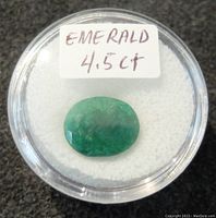 Oval-cut green emerald gemstone displayed in a round clear container with a white padded bottom and a handwritten label indicating 'EMERALD 4.5 CT'. The gemstone has visible inclusions and surface irregularities.