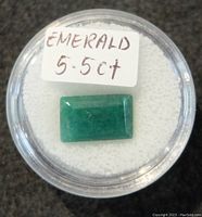 Top-down clear view of a rectangular green emerald gemstone in a clear round display case with white padding inside and a handwritten label showing 'EMERALD 5.5CT'