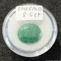 Top view of the 8.5CT emerald gemstone in a round clear case with white background, showing green tones and inclusions.