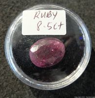 Oval faceted ruby gemstone weighing 8.5 carats, housed in a clear round container with a handwritten label.