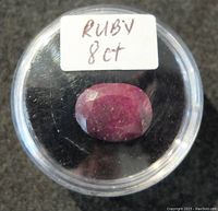 Top view of the 8 carat oval-shaped ruby gemstone inside a clear round plastic container with label 'RUBY 8 ct'. The gemstone shows visible natural inclusions.