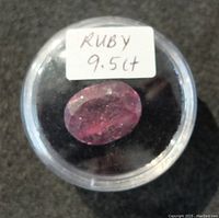 Clear plastic round display case holding an oval-shaped ruby gemstone with a white label stating 'RUBY 9.5 ct'. The ruby shows a faceted cut with visible inclusions and some surface scratches.