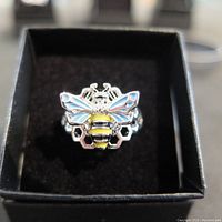 Top view of silver bee ring with yellow and black enamel on the bee body, blue enamel on wings, and honeycomb pattern on band inside a black box.