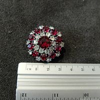 Front view of the brooch showing round blood pigeon red and clear diamond-type stones with a ruler for size reference.
