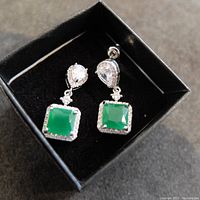 Close-up photo of the earrings showing the square green stones surrounded by clear stone halos, with pear-shaped clear stones at the posts, set in silver-tone metal and displayed in a black box.