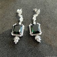 Front view of pair of emerald green square-cut stones surrounded by a halo of small clear stones, with additional clear stones above and below in a drop style, silver-tone metal setting.