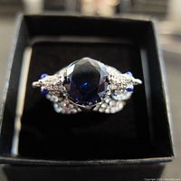 Close-up front view of ring showing the large oval royal blue sapphire center stone and surrounding diamond accents on the bird design.