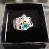 Front view of the wide silver ring with textured surface and two blue topaz-type stones in bezel settings, displayed in a black ring box.