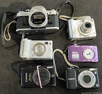 Top view showing Canon AE-1 35mm camera body, various digital cameras including Nikon Coolpix purple, Canon PowerShots in black and silver, and Fujifilm FinePix silver model