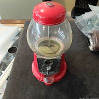 Front view of the vintage glass bubble gum machine showing red metal lid and base, clear glass globe, black coin mechanism, and small plastic catch tray.