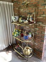Full view of the brass shelf displaying all three glass shelves and various decorative items placed on them, positioned next to a brick wall and curtain.