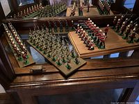 Multiple rows of painted lead toy soldiers arranged on wooden surface with mirror reflection, includes redcoat and khaki figures on green bases