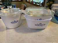 Three vintage Corningware dishes, two rectangular nested and one round, all with Blueflower pattern and clear glass lids.