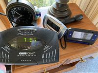 Photo showing vintage black analog clock, ObusForme sound therapy alarm clock with digital display, a silver digital alarm clock, a navy blue L.L.Bean digital alarm clock, and a black flashlight on a wooden surface.