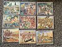 Top view of ten vintage military-related collector cards laid out in two rows on carpeted surface showing colorful historical battle scenes.