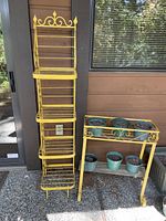 Photo of 2 yellow vintage metal plant stands against wooden wall and glass door, with some plant pots placed on the shorter stand.