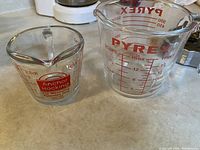 Two glass measuring cups on countertop: smaller Anchor Hocking and larger Pyrex, both with red measurement markings.