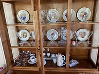 Wooden cabinet displaying the lot's dishes, glasses, and ceramics grouped on 3 shelves, showing overall contents.