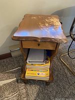 Vintage solid wood end table showing scalloped top edge, single drawer with wooden knob, lower open shelf with papers, and carved feet.