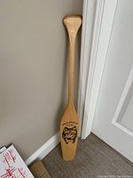 Full image of the wooden paddle leaning against a wall showing the full shape from handle to blade.