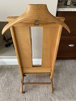 Front view of solid wood butler valet with hanger, mirror, tray, and pant holder bar on carpeted floor.