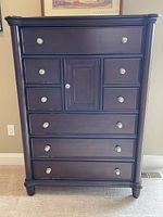 Front view of the tall chest showing dark wood finish, nine drawers, and central cabinet with silver round knobs.