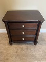 Front view of the dark brown wooden side table with three drawers and round metal knobs.