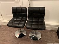 Front view of two black pedestal chairs with black faux leather tufted seats and backs, chrome pedestal bases on wooden floor.