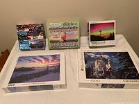 Five puzzles displayed on a table with box artwork showing various scenic and thematic images including castle, sunset, train, and people.