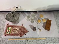 Antique galvanized metal bread maker with crank lid alongside four glass jars, a wooden box, a small metal spice grinder, a small spoon, and a decorative woven pouch.