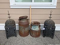 Front view of the 2 rusty antique milk cans and the 2 black metal Arabic-style lanterns arranged side by side on the ground in front of a beige wall.
