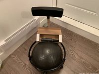 Front and side views of the ball chair showing the black exercise ball seat enclosed in a black metal ring with four legs on wheels and a black padded backrest. Manual pump in white box is attached to the metal support.