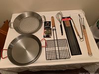 Overall view of the set including two pans, bbq skewers, folding grill basket, tongs, whisk, and fork