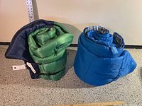 Two sleeping bags rolled up, green with blue interior on left, solid blue with plaid interior on right.