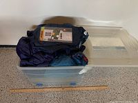 Three rolled sleeping bags inside a clear Rubbermaid container with blue wheels and a yellow lid on the floor.